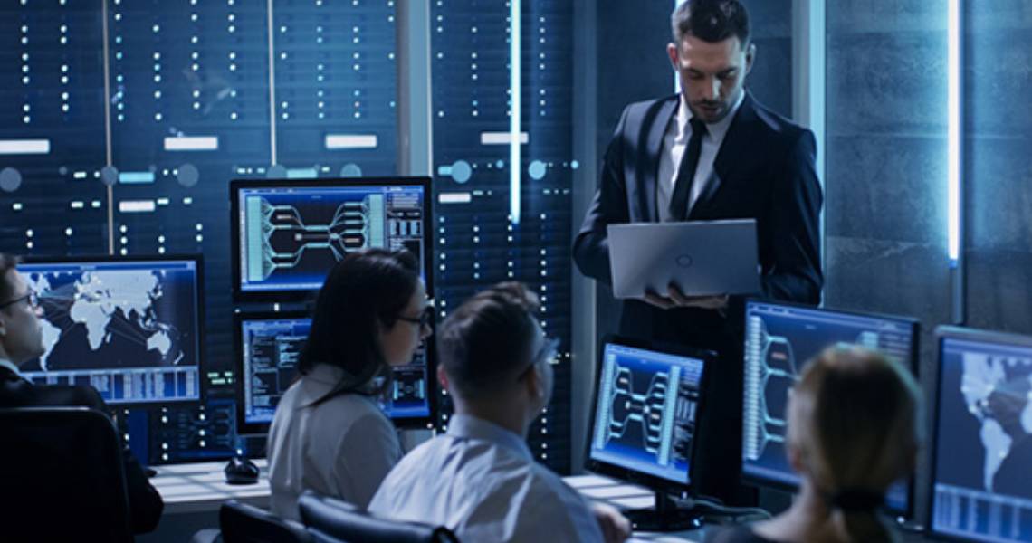 A group of colleagues working on desktops, while a man stands and works on a laptop